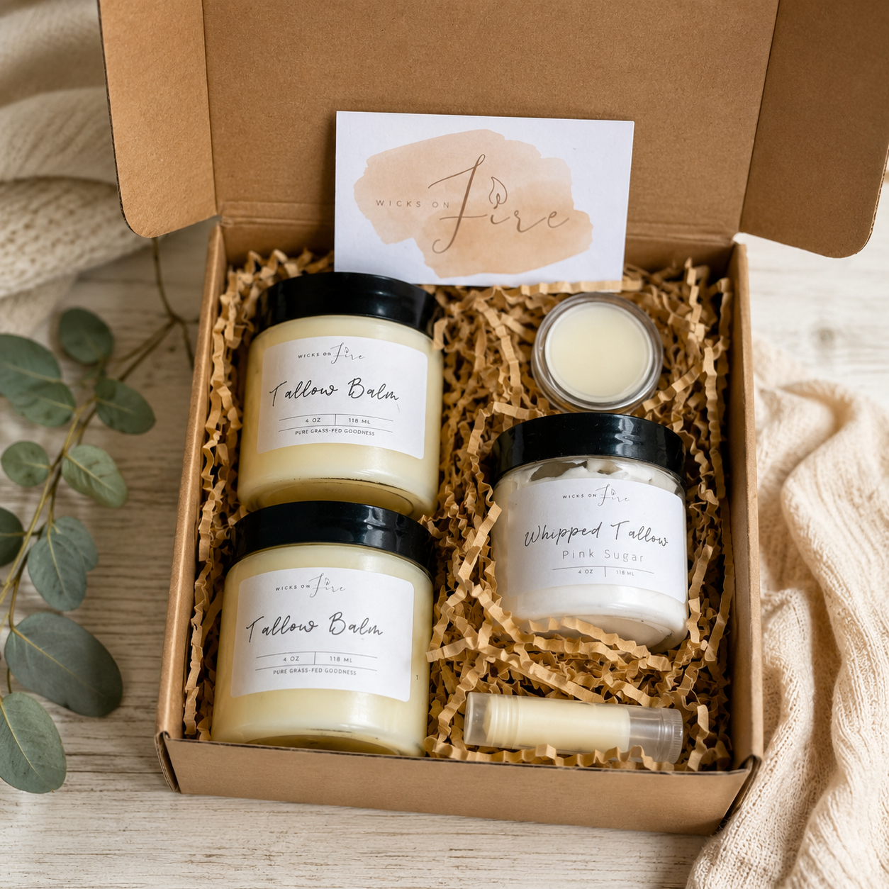 An open kraft gift box filled with wax melts and balms from the brand Wicked Fire. The set includes two 'Fallows Balm' jars, a 'Whipped Fallows' lotion melt jar, a small lip balm tube, and a Wicked Fire brand card, all arranged in brown crinkle paper shred on a light wood surface with a beige knit blanket and eucalyptus leaves.