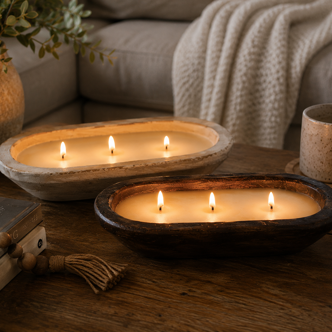 Two lit candles in rustic oval wooden and stone bowls placed on a wooden table in a cozy living room setting with a sofa, throw blanket, mug, and books in the background.