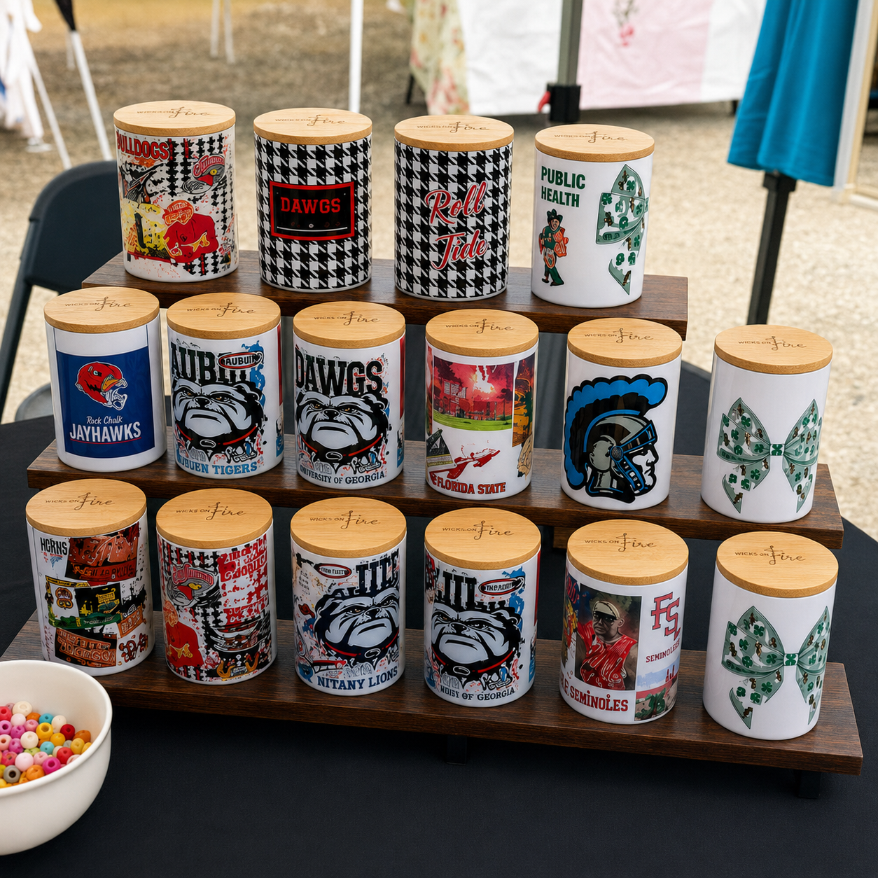A set of 16 white cylindrical storage jars with light wood lids, arranged on a wooden display stand. Each jar is decorated with a different Alabama, Auburn, Georgia sports logo, mascot, or themed graphic.