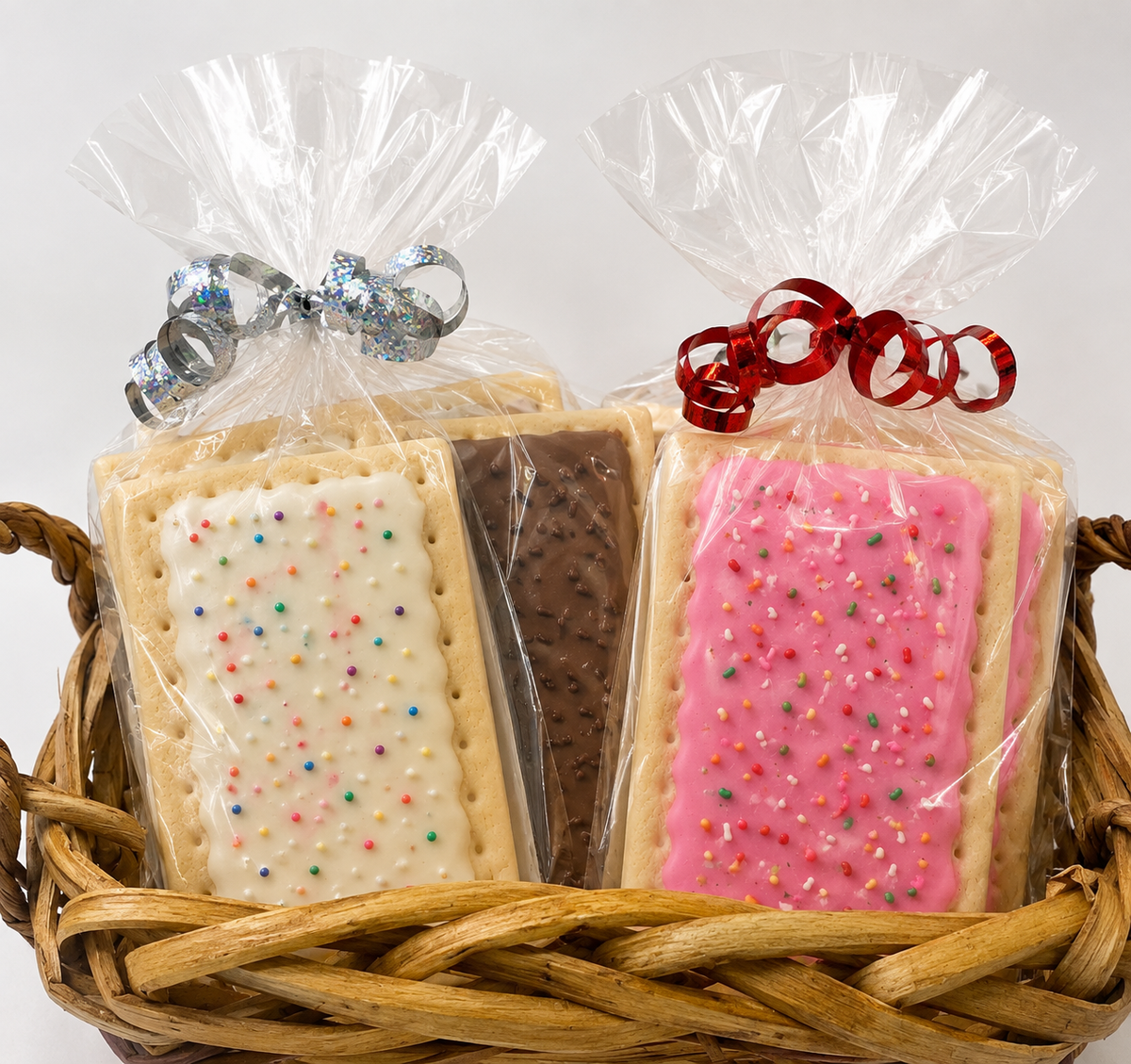 Two packaged rectangular cookies in clear cellophane bags tied with silver and red ribbons, displayed in a woven basket. One cookie is white with multicolored sprinkles and a brown cookie beside it.
